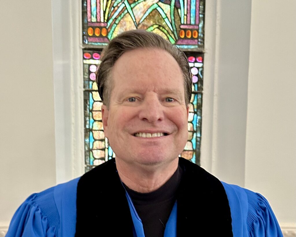 Smiling man in blue robe before stained glass