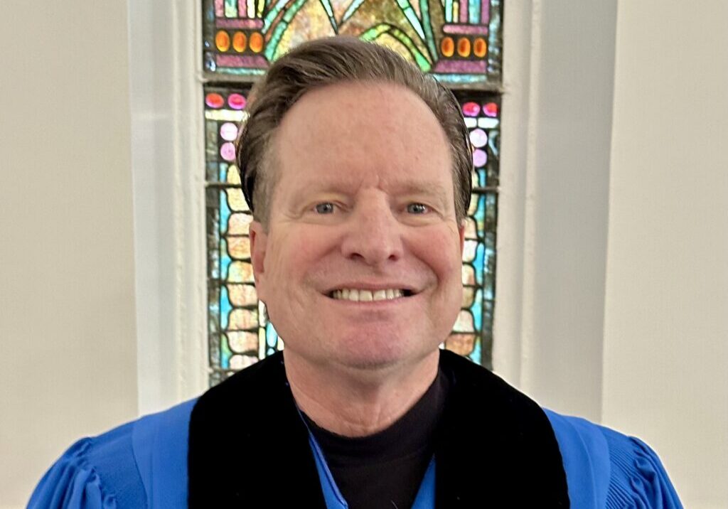 Smiling man in blue robe before stained glass